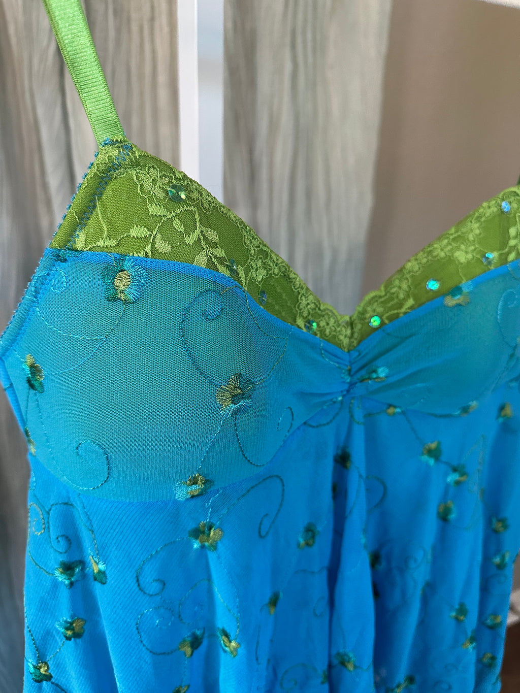 A close-up of a sheer blue lingerie chemise with green floral embroidery and sequins, featuring a lime green lace-trimmed underwire cup and a matching green shoulder strap, shown against a neutral indoor background.