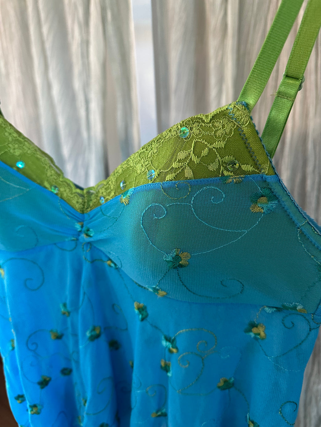 A close-up of a blue and green embroidered lingerie chemise or nightgown, featuring a sheer blue body with gold embroidery, a lime green lace-trimmed bust, and green straps, shown against a soft white curtain background.