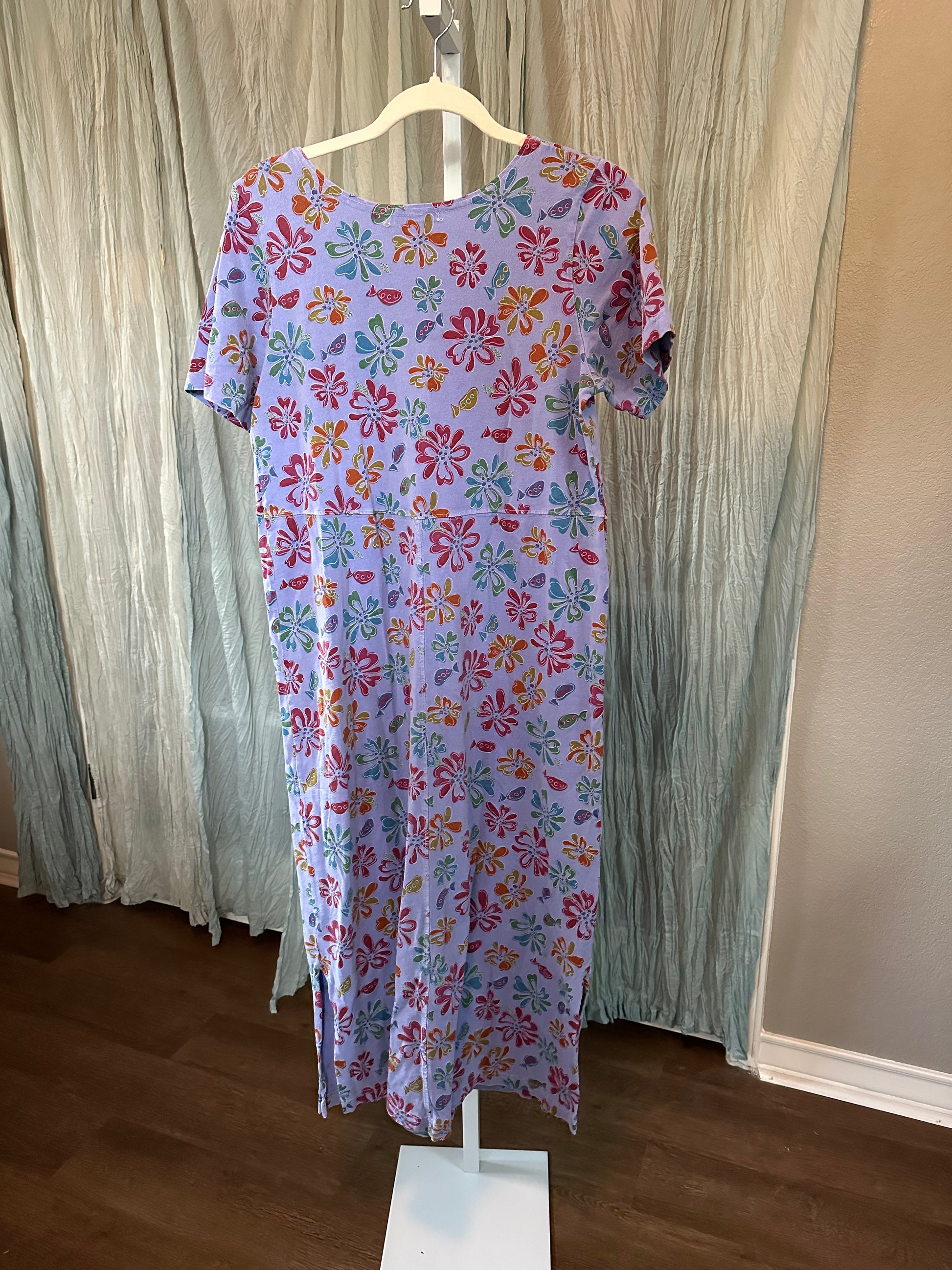 A short-sleeve midi dress with a colorful floral pattern on a light lavender background, displayed on a white stand in front of a textured sage green curtain.
