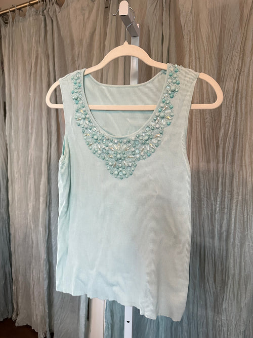 A light mint green sleeveless top with a heavily embellished beaded neckline, hanging on a wooden hanger against a gray curtain backdrop.