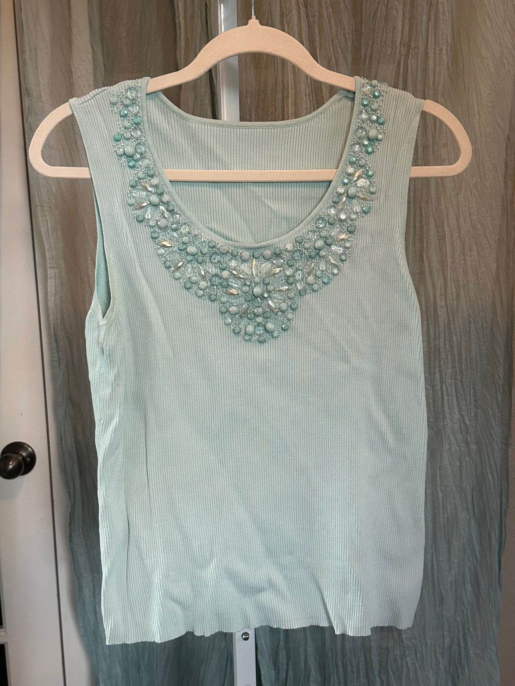 A light mint green ribbed knit sleeveless top with a decorative beaded neckline, hanging on a wooden hanger against a gray curtain background.