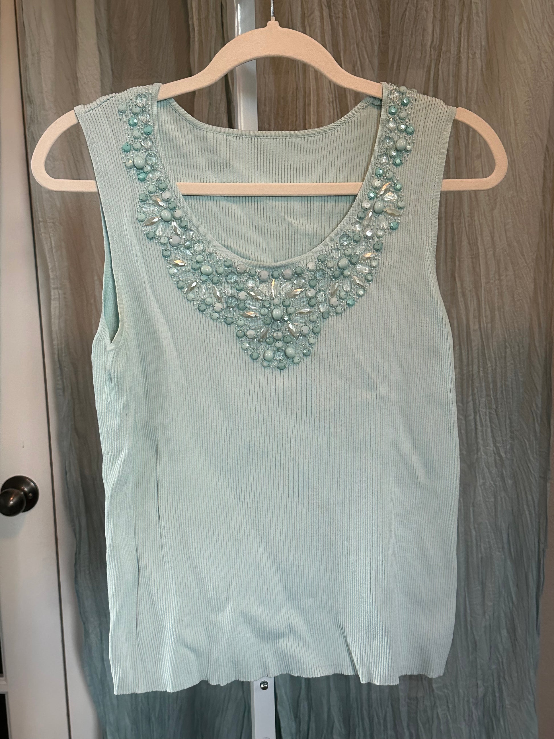 A light mint green ribbed knit sleeveless top with a decorative beaded neckline, hanging on a wooden hanger against a gray curtain background.