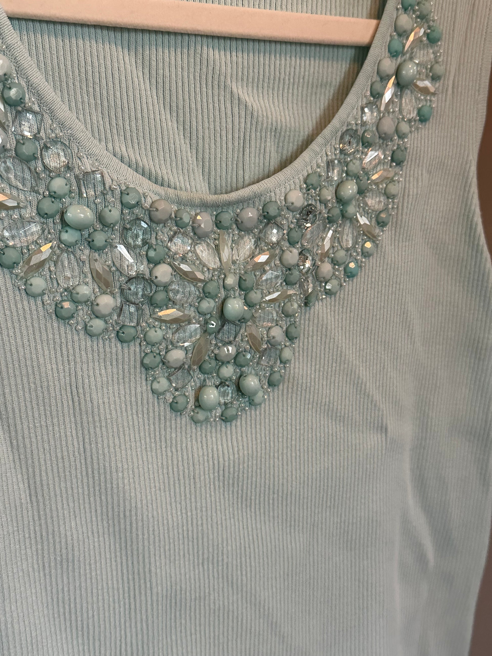 A close-up of a ribbed sage green sleeveless top on a wooden hanger, featuring a wide scoop neckline embellished with a dense arrangement of light blue-green beads and clear rhinestones.