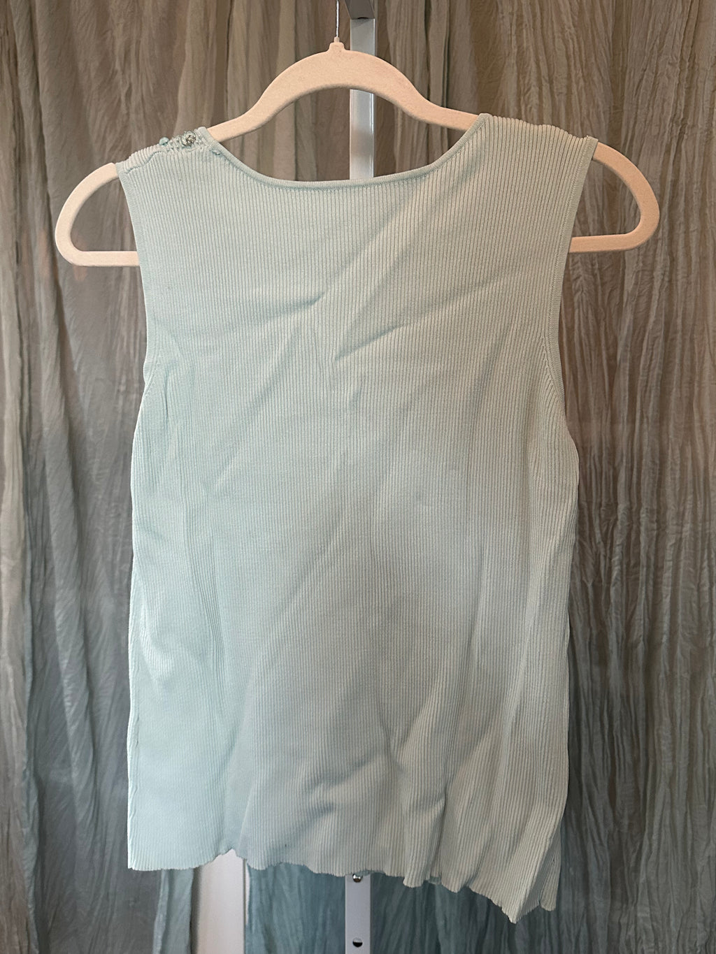 A light sage green sleeveless top with a subtle vertical ribbed texture, hanging on a white plastic hanger against a wrinkled gray curtain backdrop.