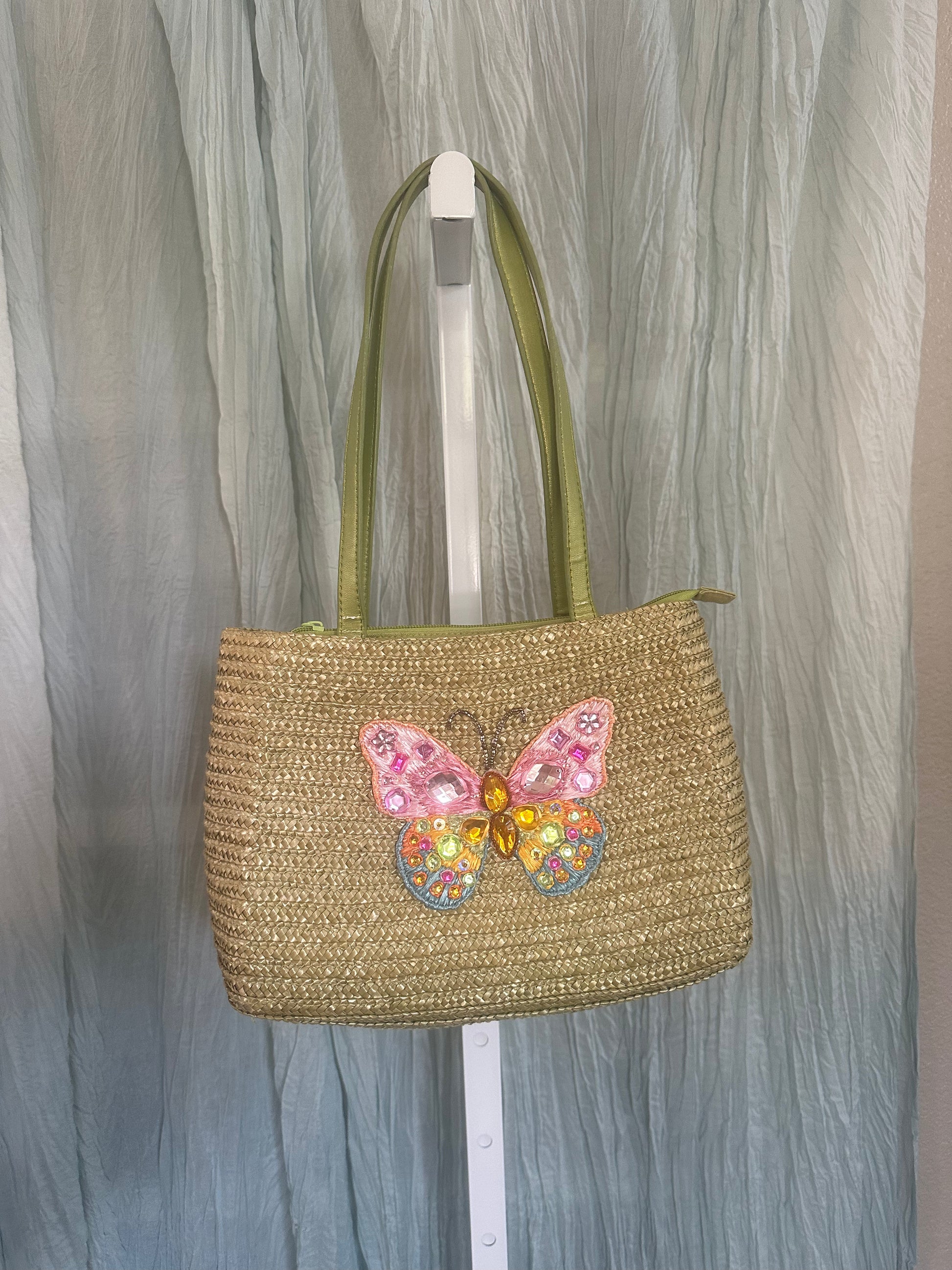 A woven straw tote bag with olive green handles and a colorful butterfly embellishment on the front, hanging on a white stand against a gray draped fabric backdrop.