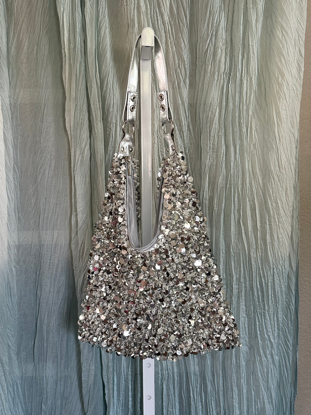 A silver sequined shoulder bag with a wide strap, hanging against a crinkled light blue-gray fabric backdrop.