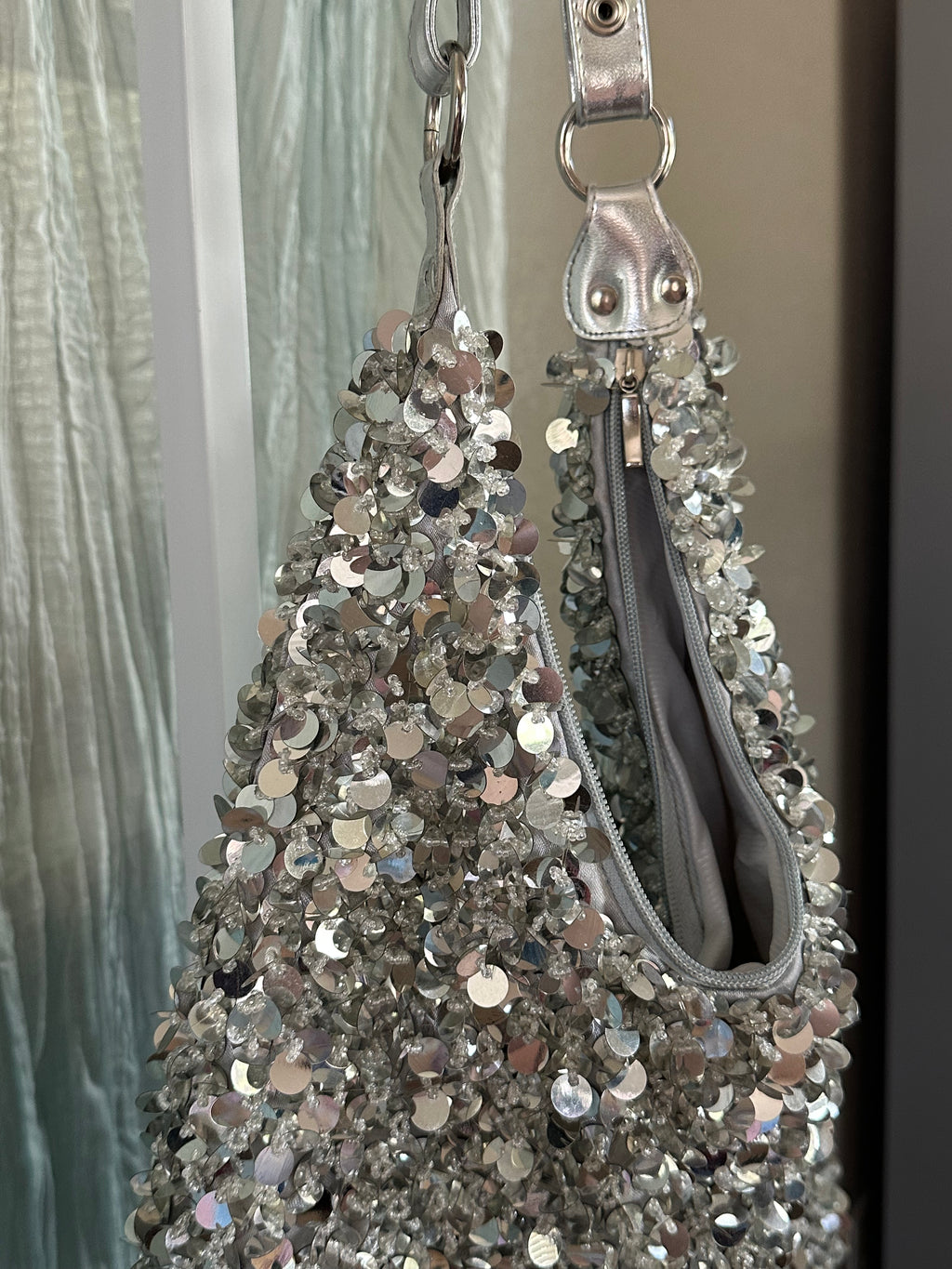 A close-up of a silver sequined handbag hanging against a curtain and wall background. The bag features a textured surface covered in reflective silver sequins, a silver zipper, and a silver metal ring handle.