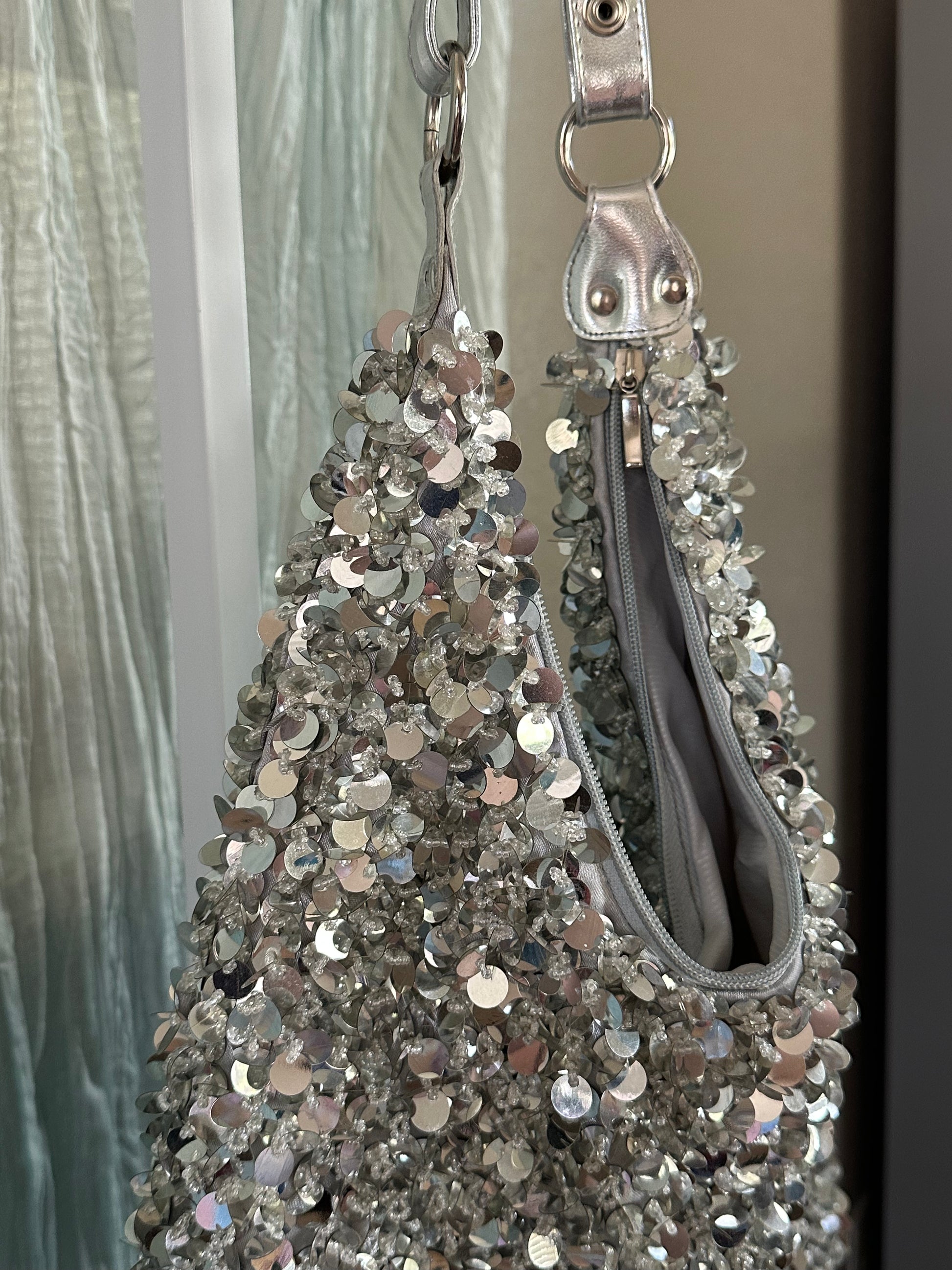 A close-up of a silver sequined handbag hanging against a curtain and wall background. The bag features a textured surface covered in reflective silver sequins, a silver zipper, and a silver metal ring handle.