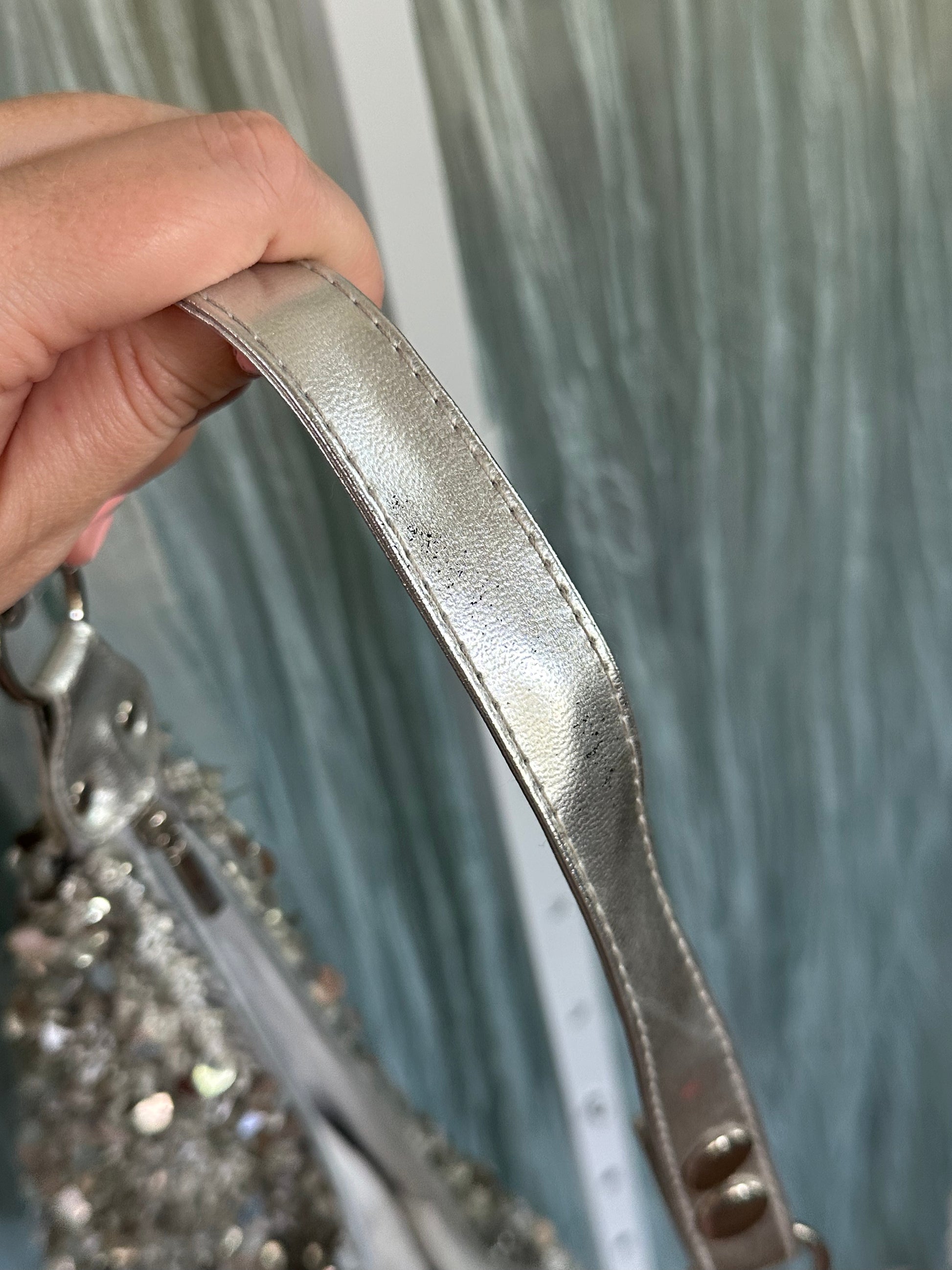 A close-up of a hand holding a silver metallic leather strap of a embellished handbag against a blue-gray curtain background.