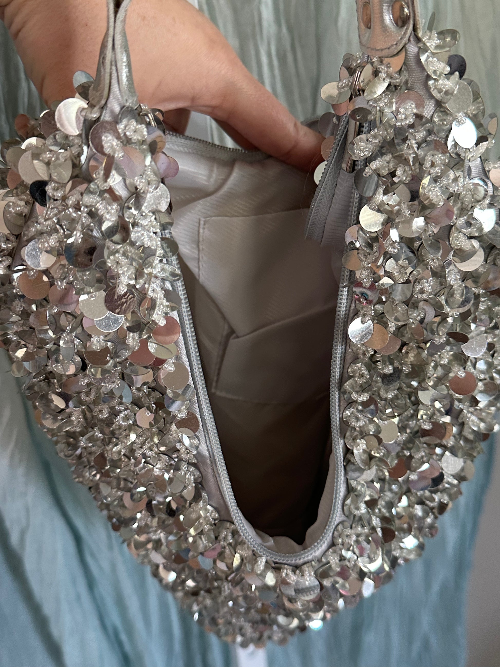 A close-up of a hand holding an open silver sequined handbag, showing the interior lining and zipper, against a light blue fabric background.