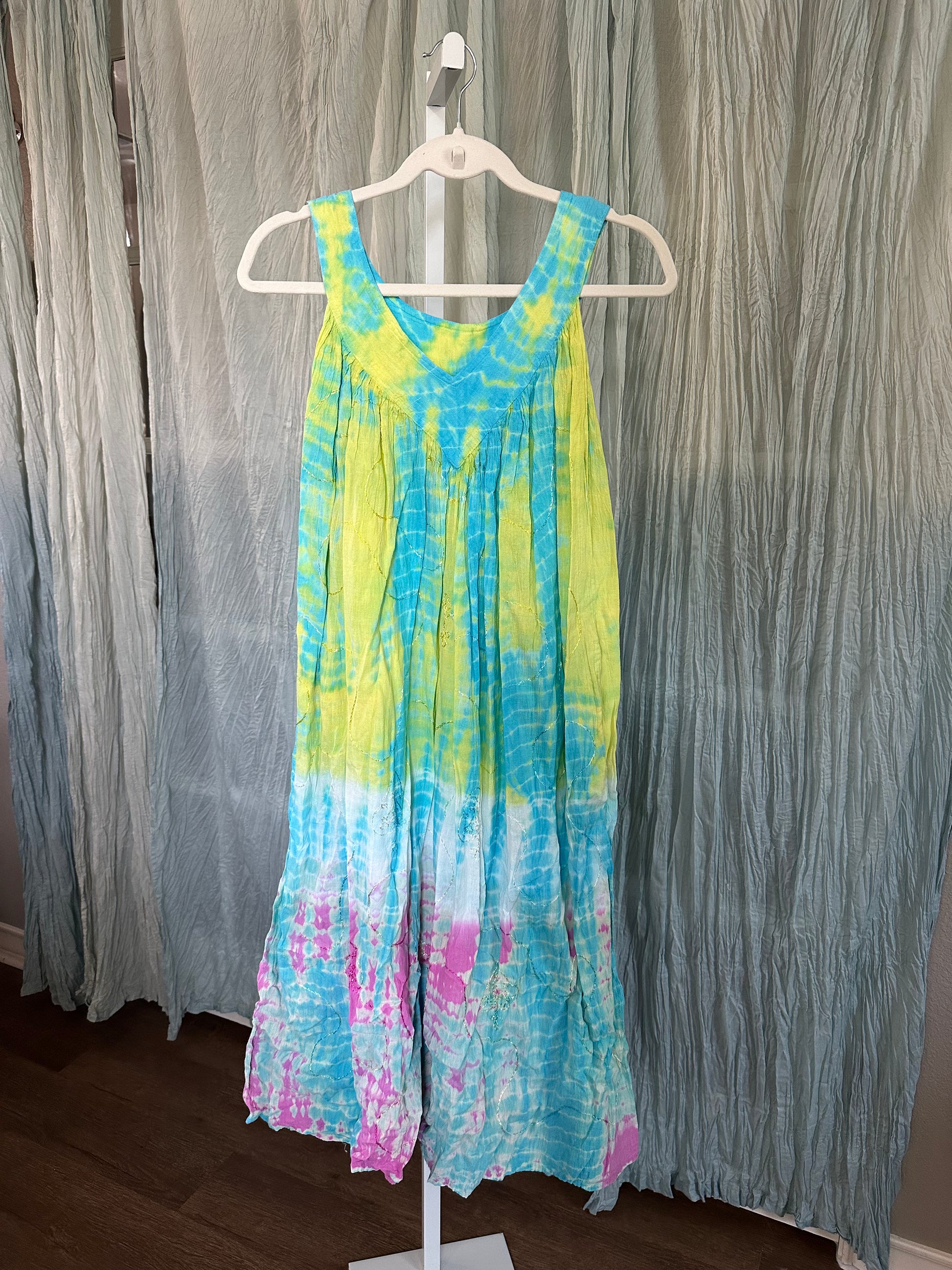 A sleeveless tie-dye jumpsuit in yellow, blue, white, and pink, hanging on a white hanger against a blue-gray curtain backdrop.