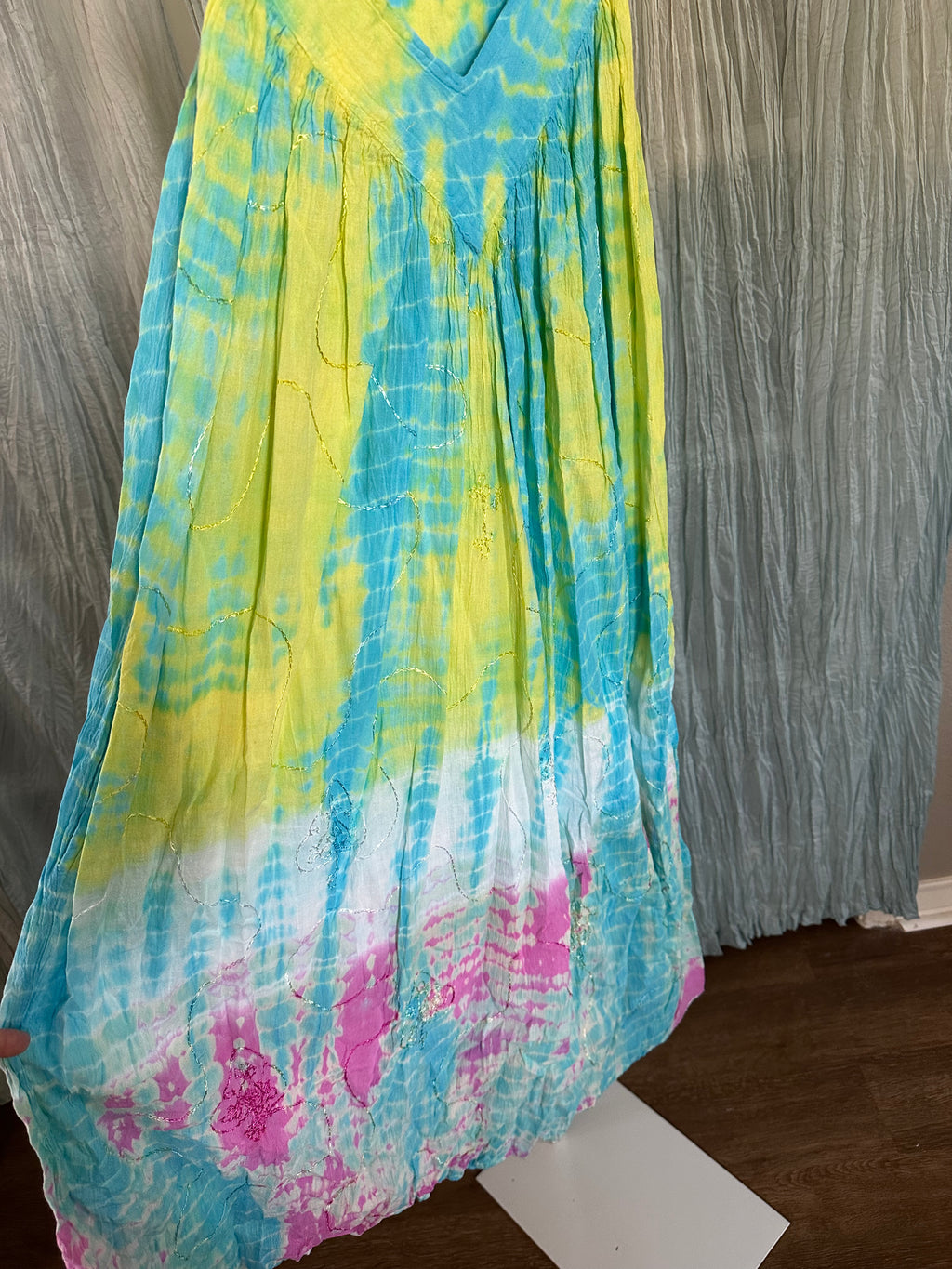 A sleeveless V-neck maxi dress with a vibrant tie-dye pattern in yellow, blue, white, and pink, displayed against a gray curtain backdrop.