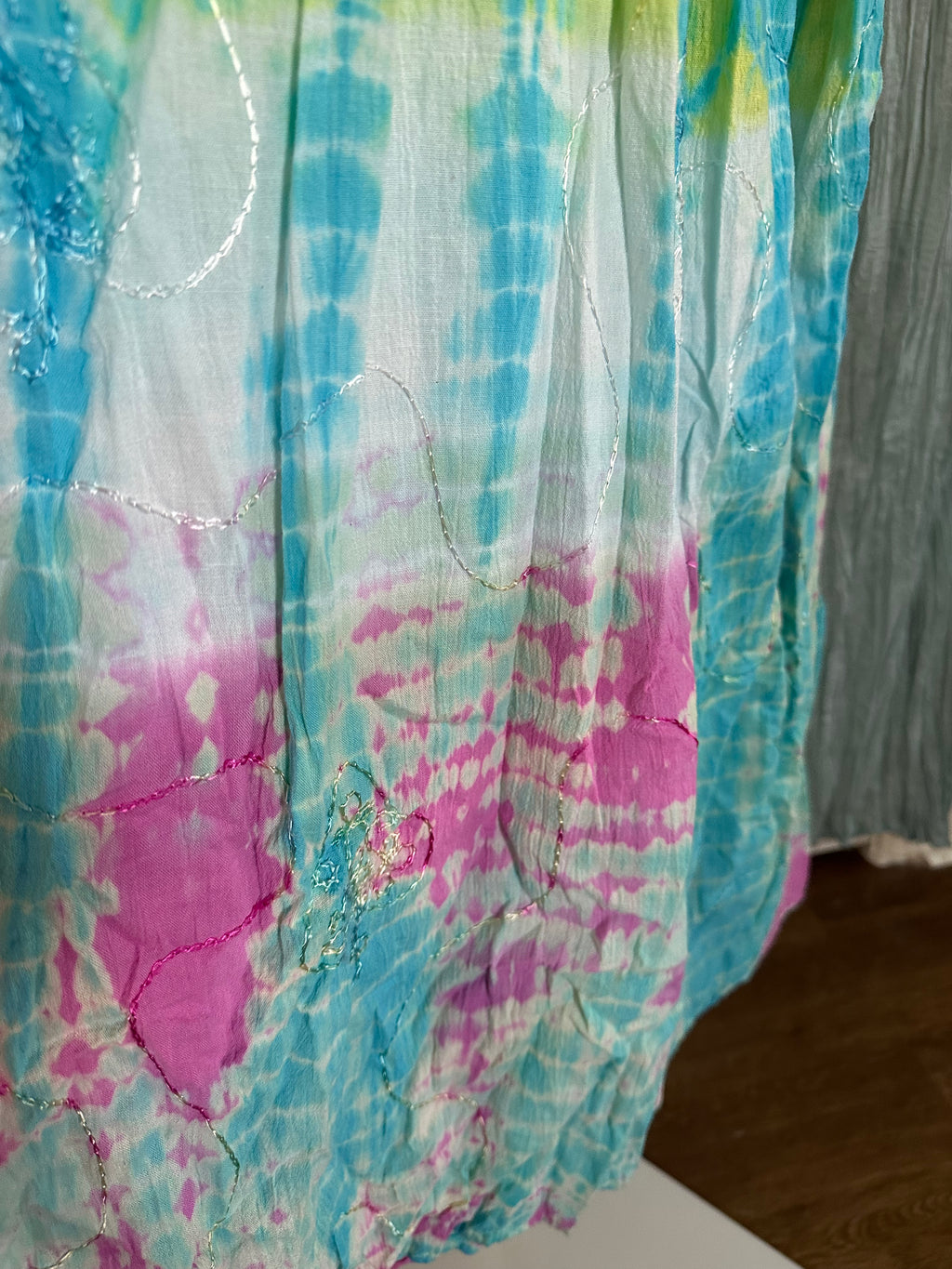 A close-up of a lightweight fabric with a tie-dye pattern in turquoise blue, pink, and white, featuring subtle metallic embroidered lines.