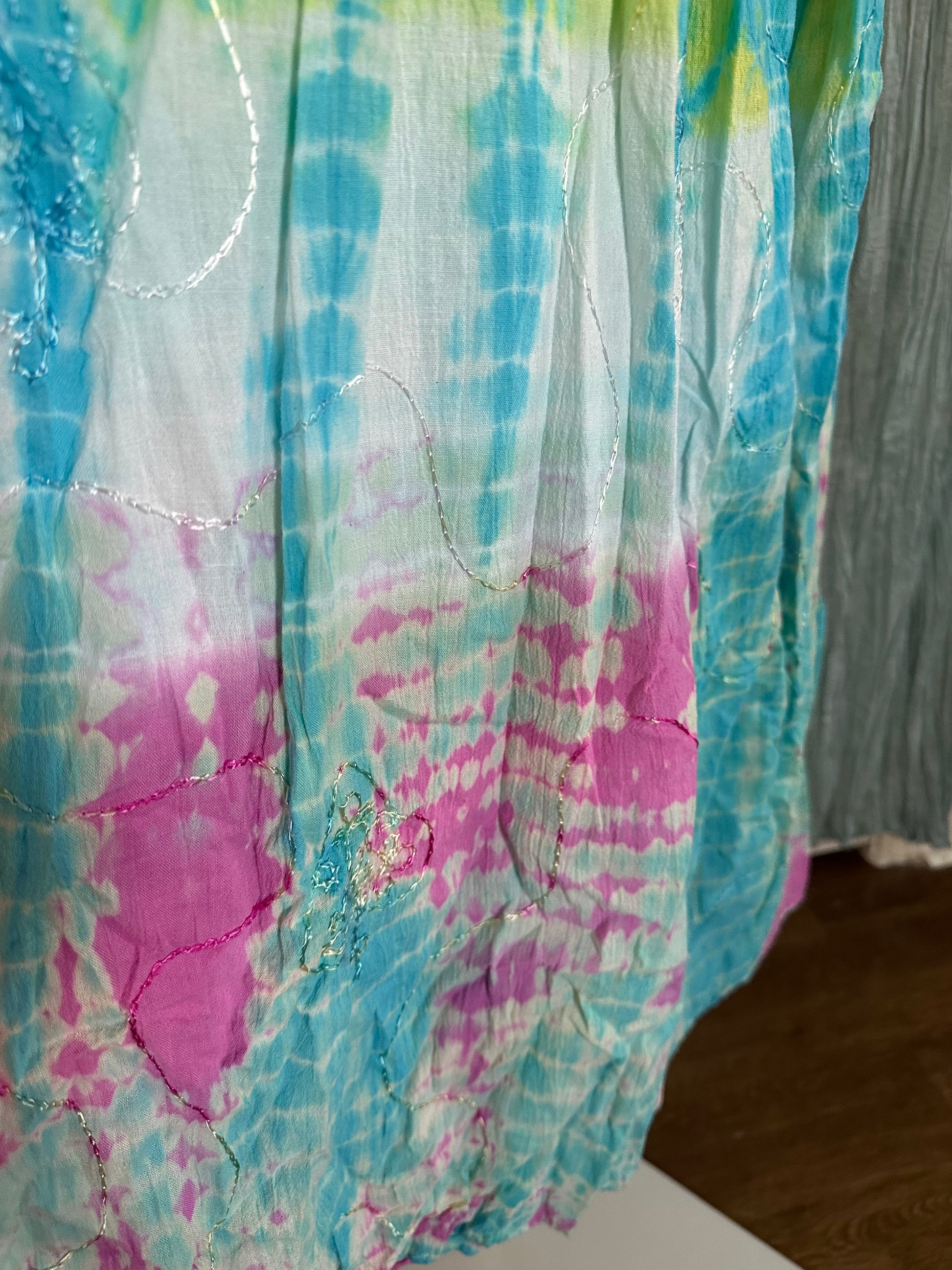 A close-up of a lightweight fabric with a tie-dye pattern in turquoise blue, pink, and white, featuring subtle metallic embroidered lines.