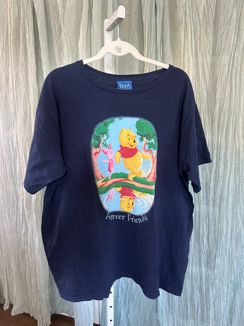 A navy blue short-sleeve T-shirt hanging on a white plastic hanger against a light gray curtain backdrop. The shirt features a colorful front graphic of Winnie the Pooh and his friends in an oval scene with the text 'Forever Friends' below.