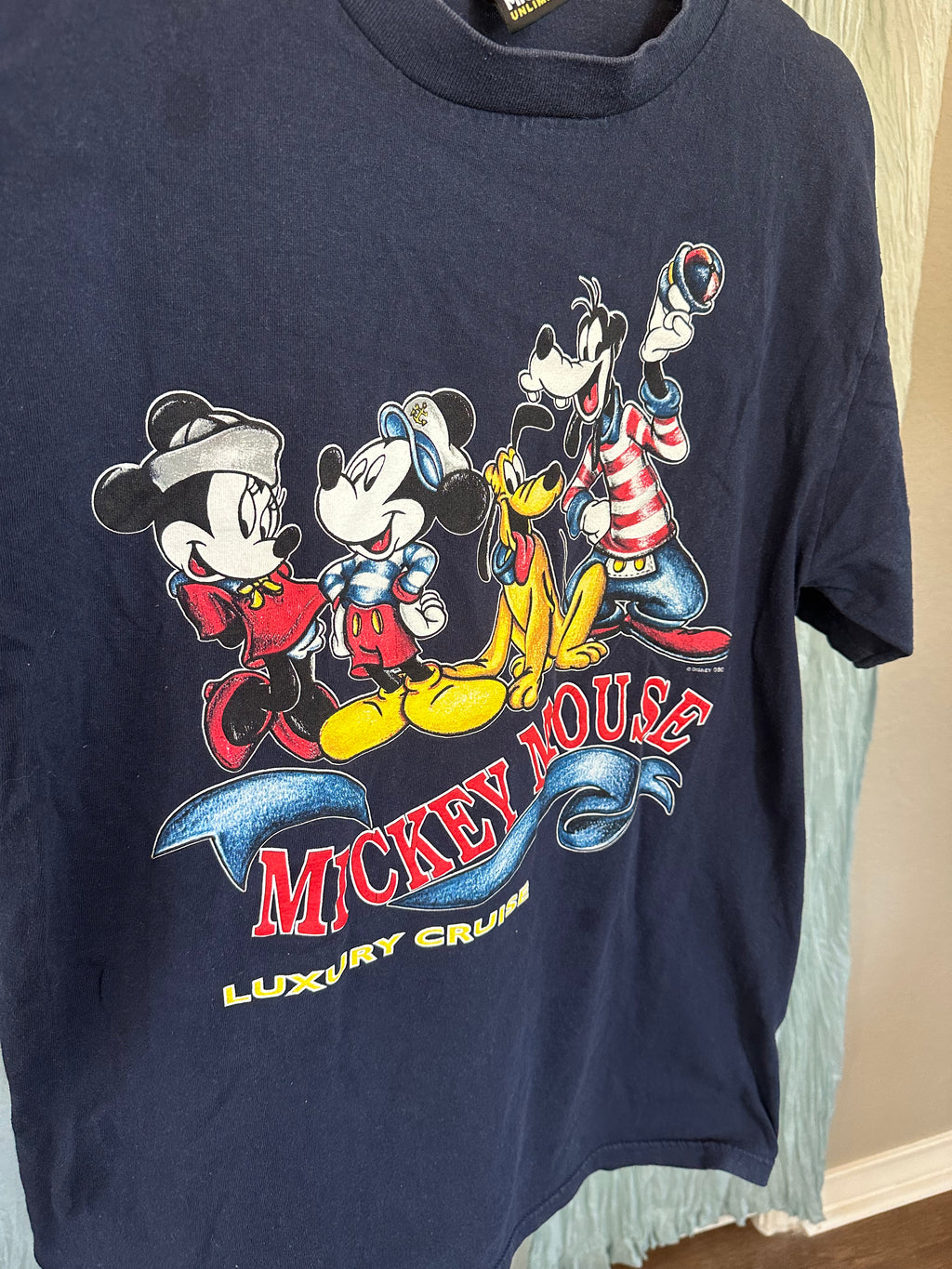 A navy blue Mickey Mouse graphic t-shirt hanging on a light blue garment hanger. The front features a colorful print of Mickey, Minnie, Goofy, and Pluto with the text 'MICKEY MOUSE LUXURY CRUISE'.