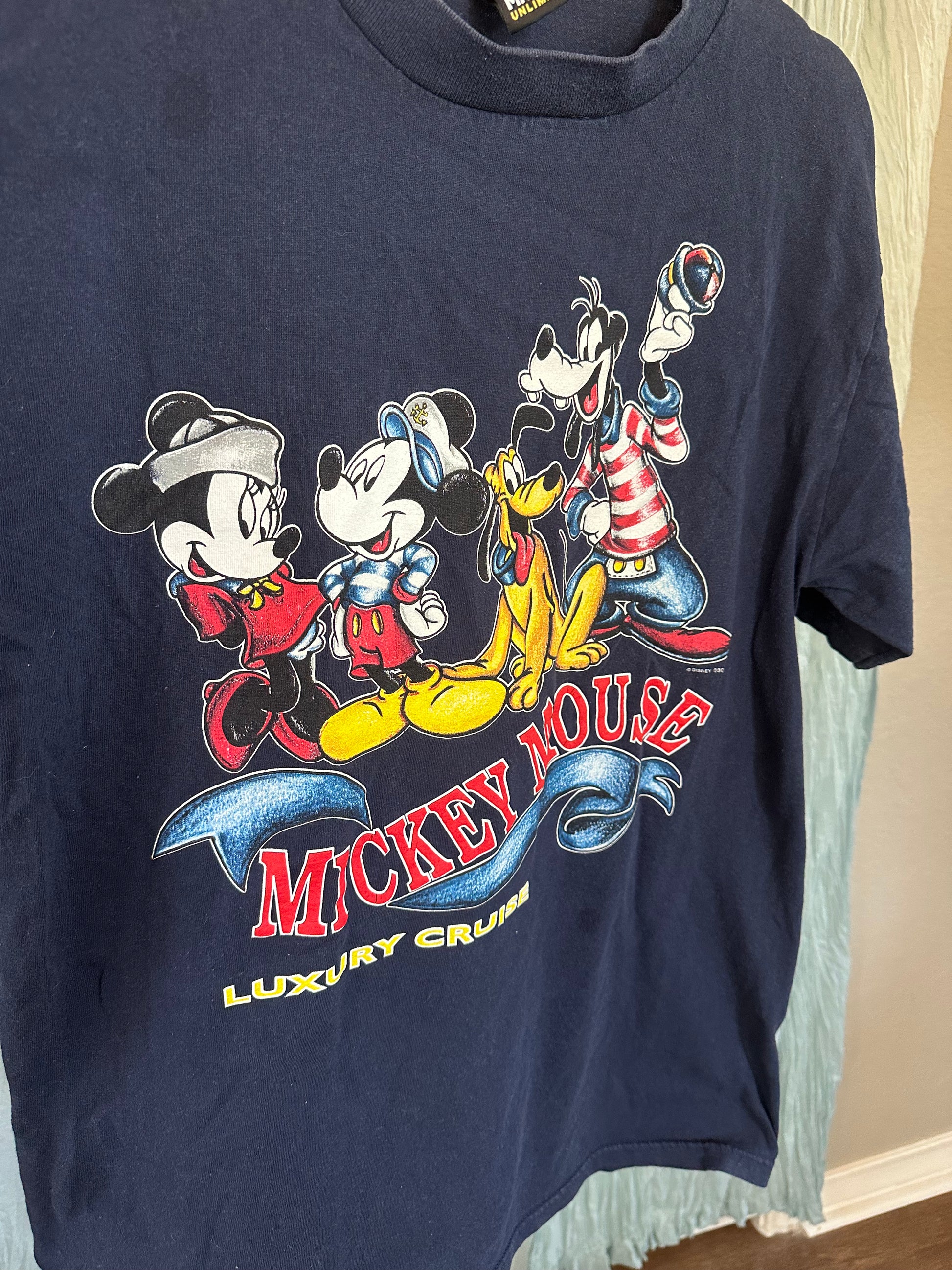 A navy blue Mickey Mouse graphic t-shirt hanging on a light blue garment hanger. The front features a colorful print of Mickey, Minnie, Goofy, and Pluto with the text 'MICKEY MOUSE LUXURY CRUISE'.