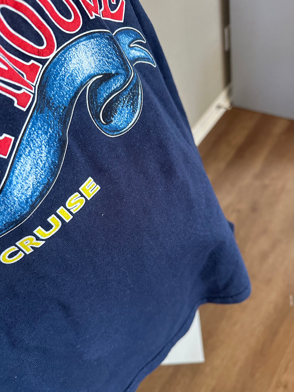 Close-up of a navy blue vintage graphic t-shirt featuring a blue snake design and yellow 'CRUISE' text, with a blurred indoor room background.