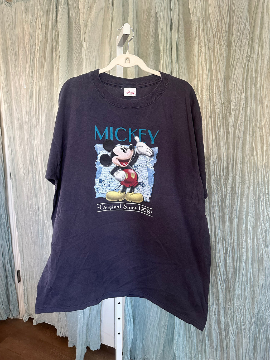 A dark navy blue Mickey Mouse graphic t-shirt hanging on a white plastic hanger against a light blue textured curtain backdrop.