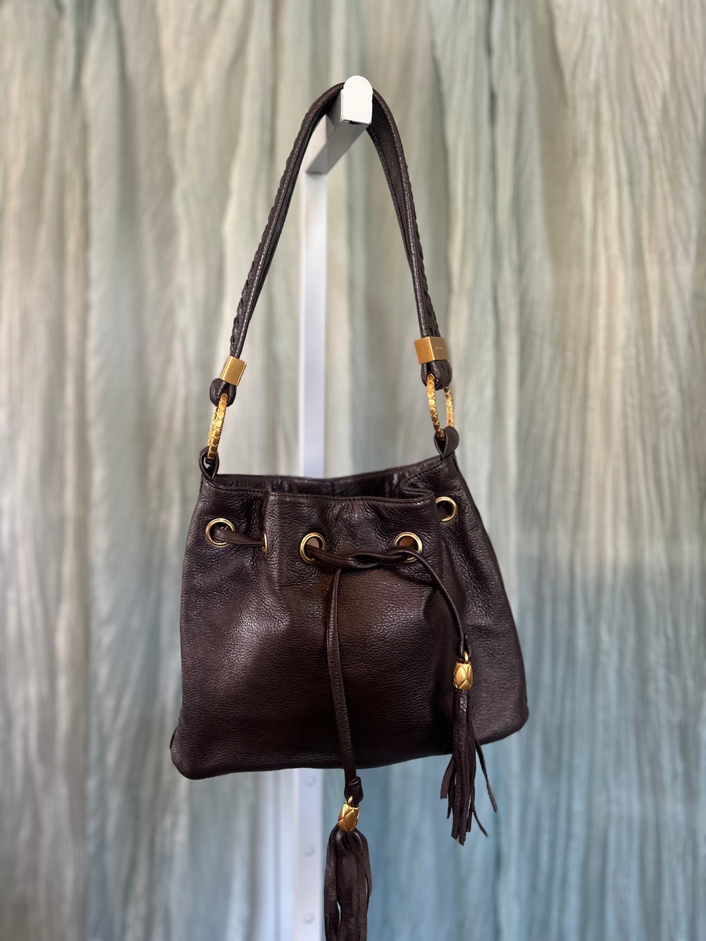 A dark brown leather drawstring bucket bag with gold-tone hardware, hanging from a white display stand against a textured gray background.