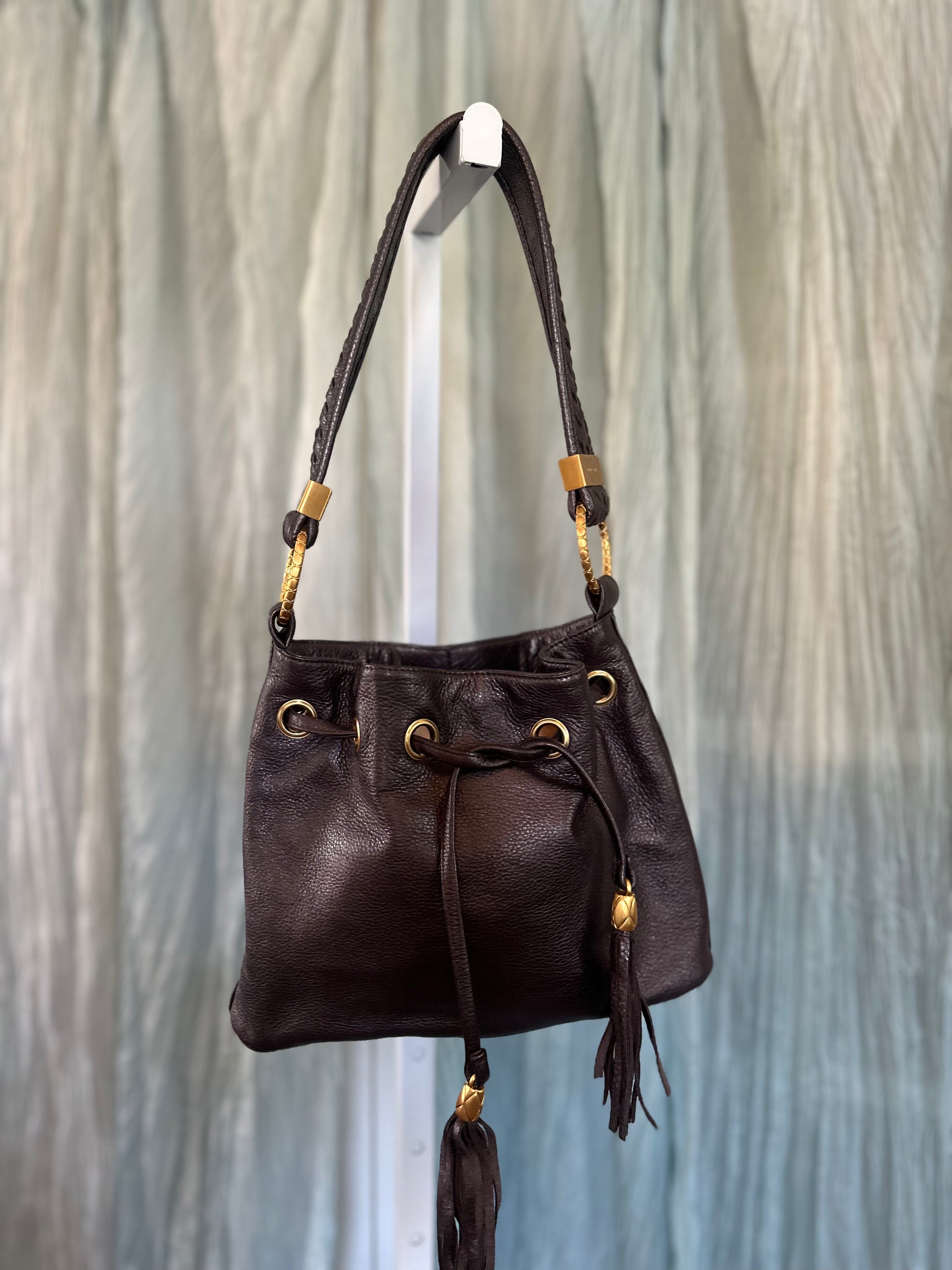 A dark brown leather drawstring bucket bag with gold-tone hardware, hanging from a white display stand against a textured gray background.
