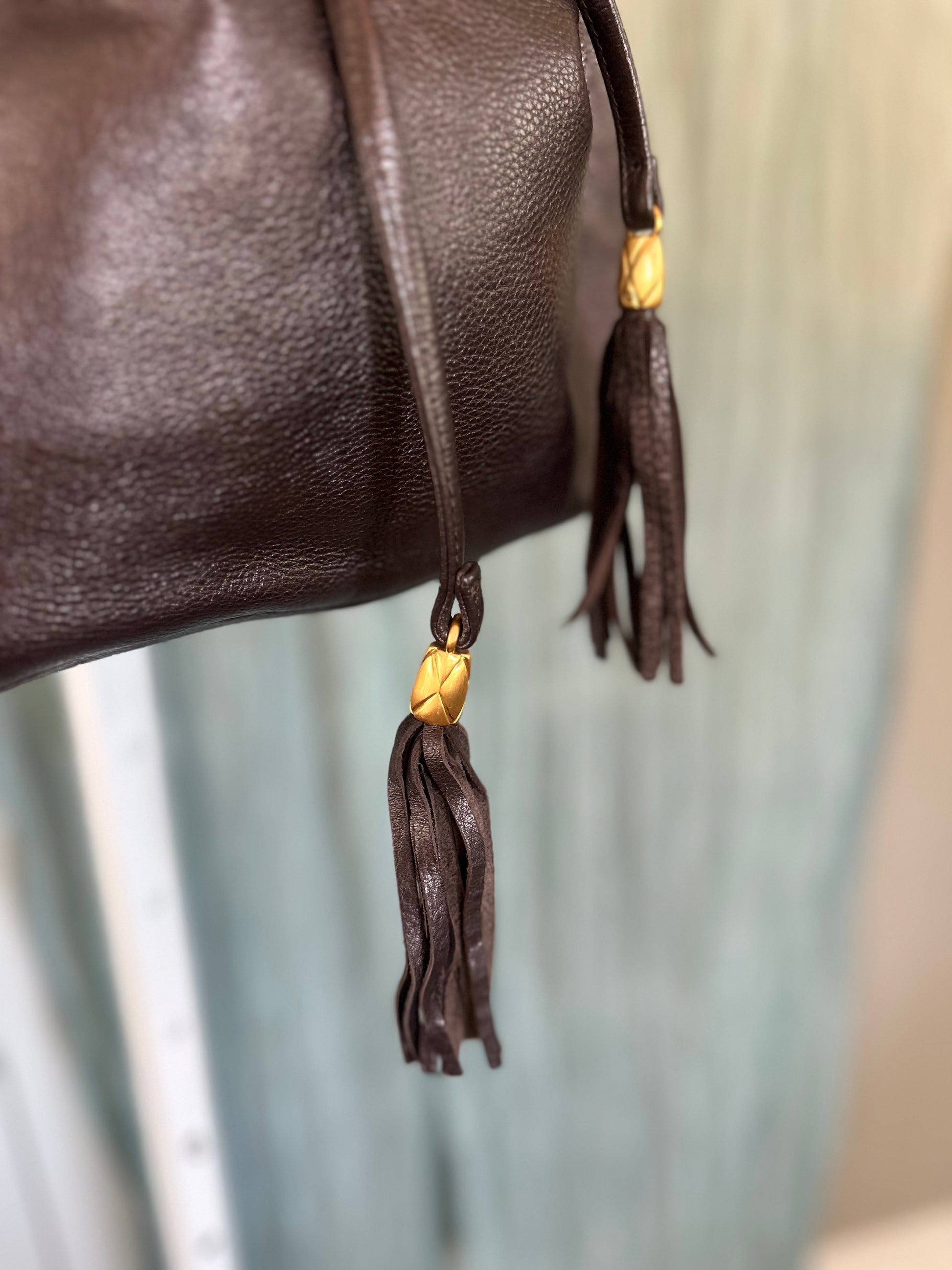 Close-up of a dark brown textured leather bag with hanging tassels and small gold-toned accents, photographed against a softly blurred light blue and beige background.