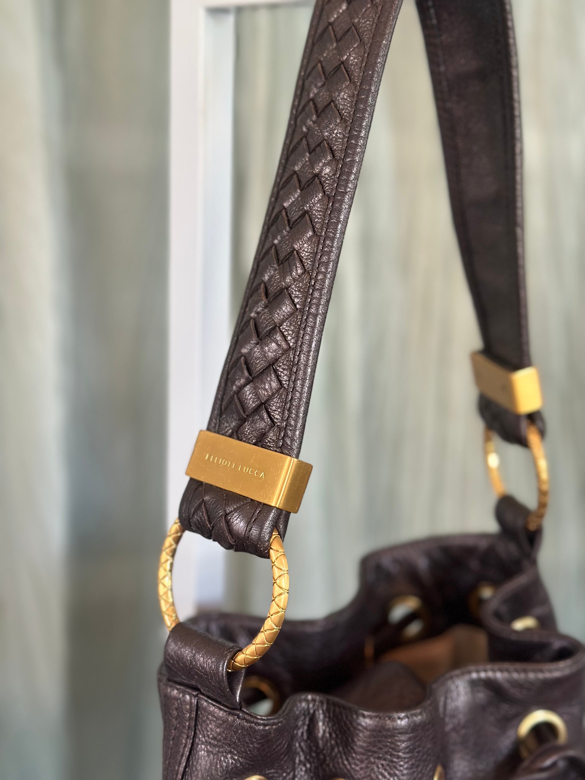 Close-up of a dark brown leather handbag with a woven shoulder strap and gold-tone hardware, including a visible rectangular brand plaque engraved 'Elliott Lucca'.