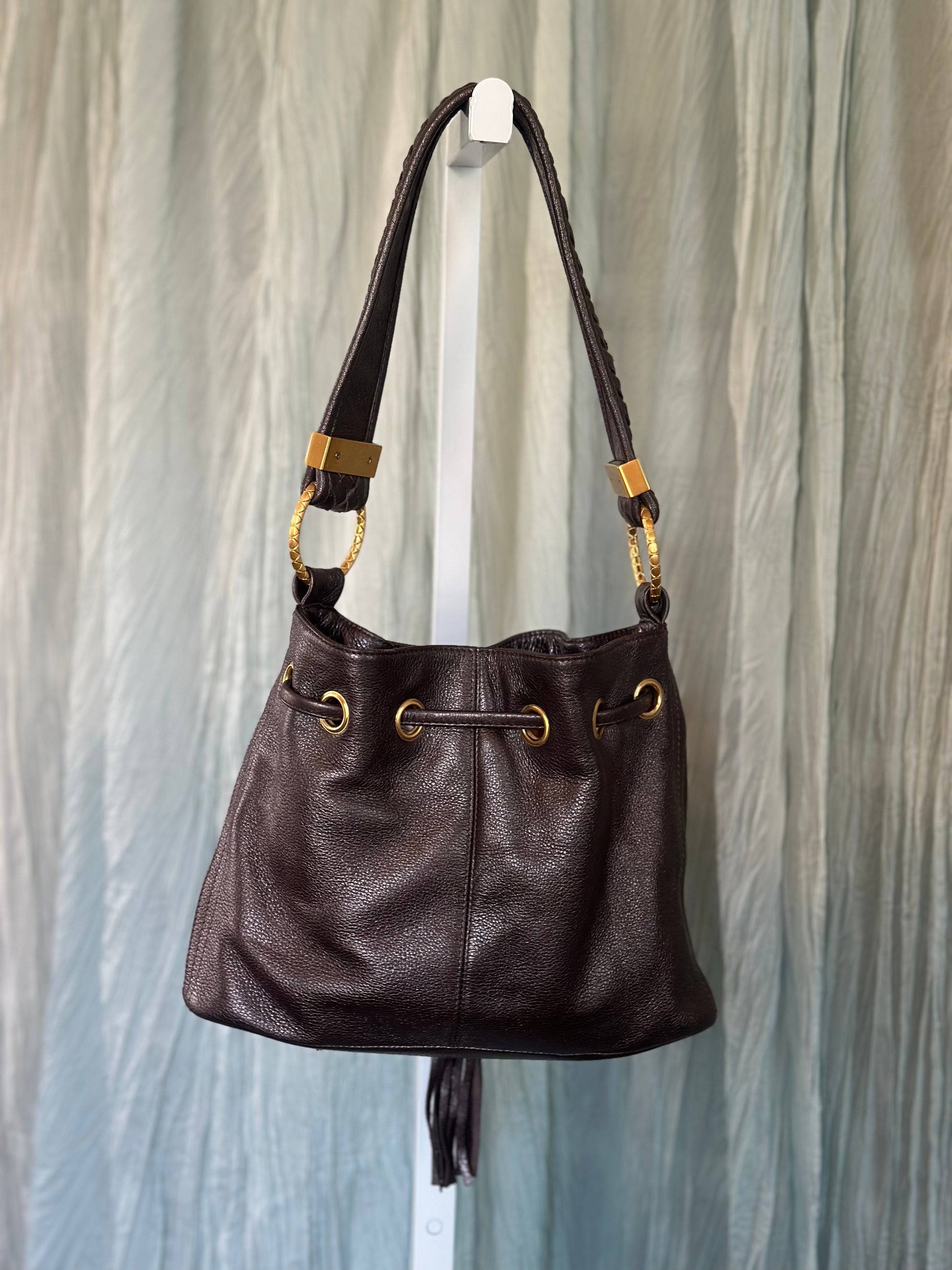 A dark brown leather shoulder bag with gold-tone hardware, hanging on a white display stand against a light blue-gray draped fabric background.