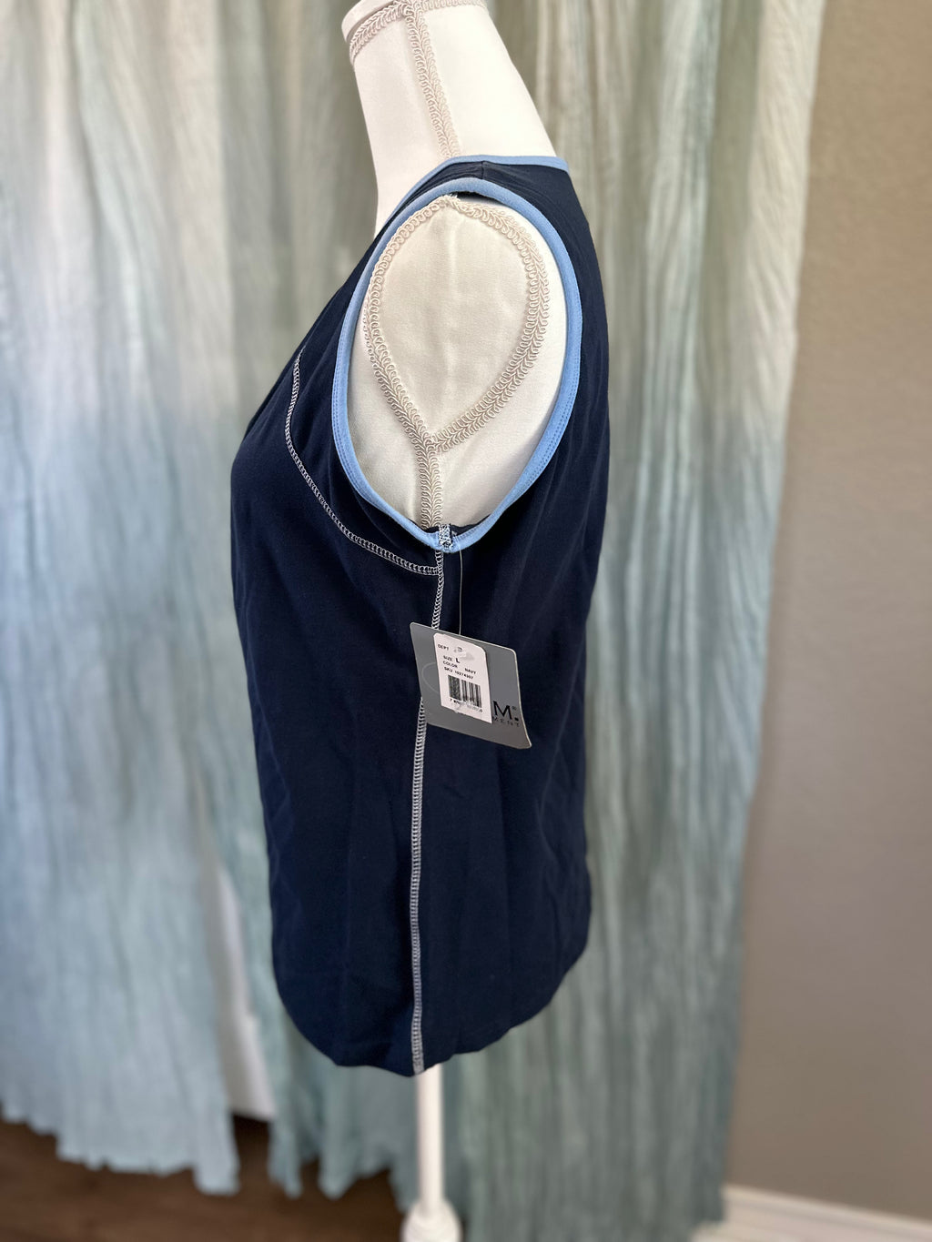 A sleeveless navy blue athletic vest displayed on a white mannequin torso, featuring light blue trim and a white padded shoulder insert. A product tag is attached near the hem, set against a background of light blue pleated fabric and a neutral wall.