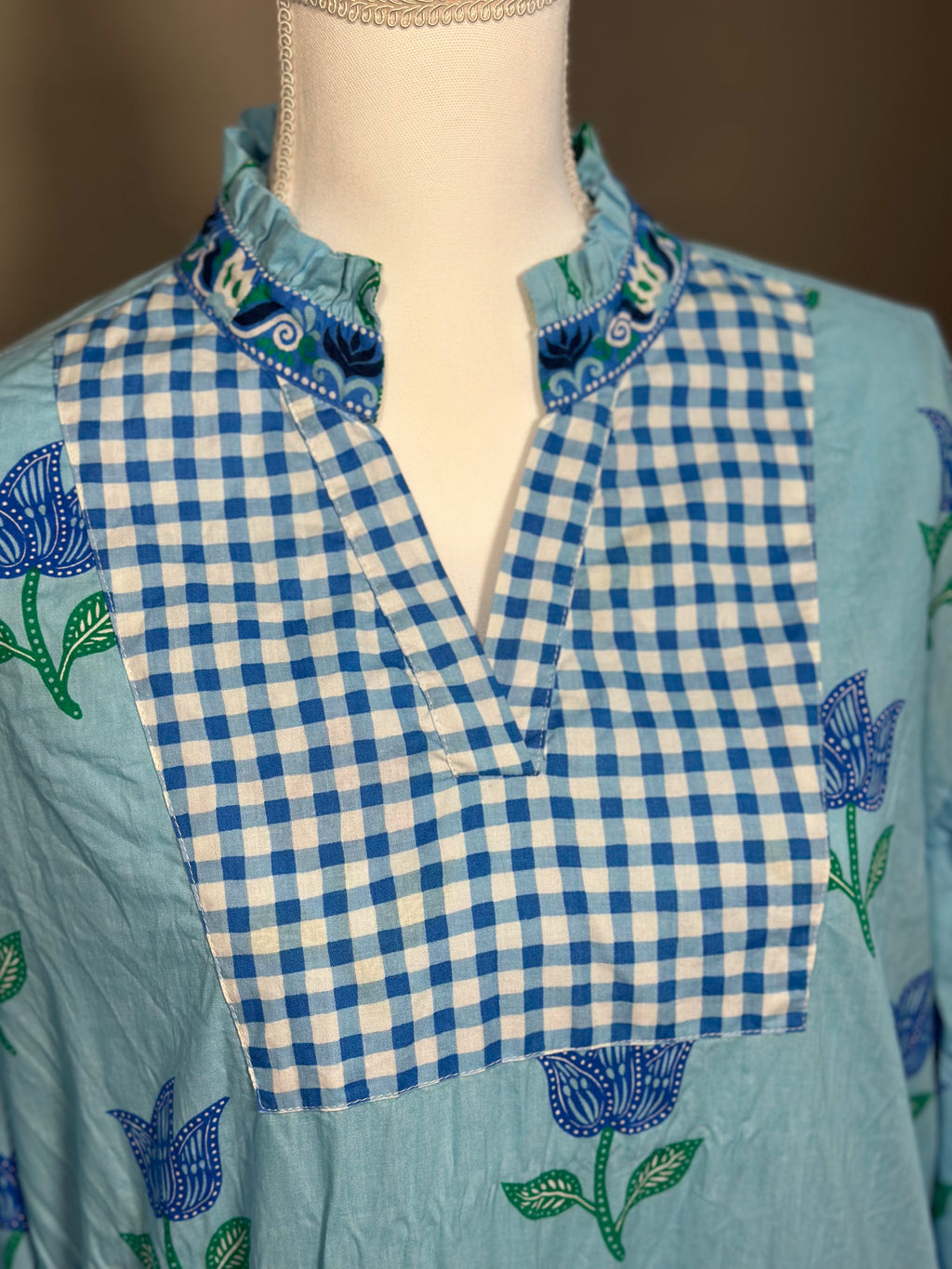 A light blue cotton tunic top with a blue and white gingham check V-neck panel, decorated with blue and green floral embroidery, displayed on a cream dress form.